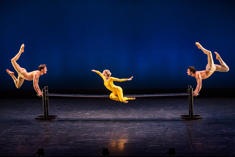 A woman in a yellow unitard sits on a wooden plank. Two men leap into the air, holding onto to each of the plank's ends. Their backs arch and their feet point to the ceiling.